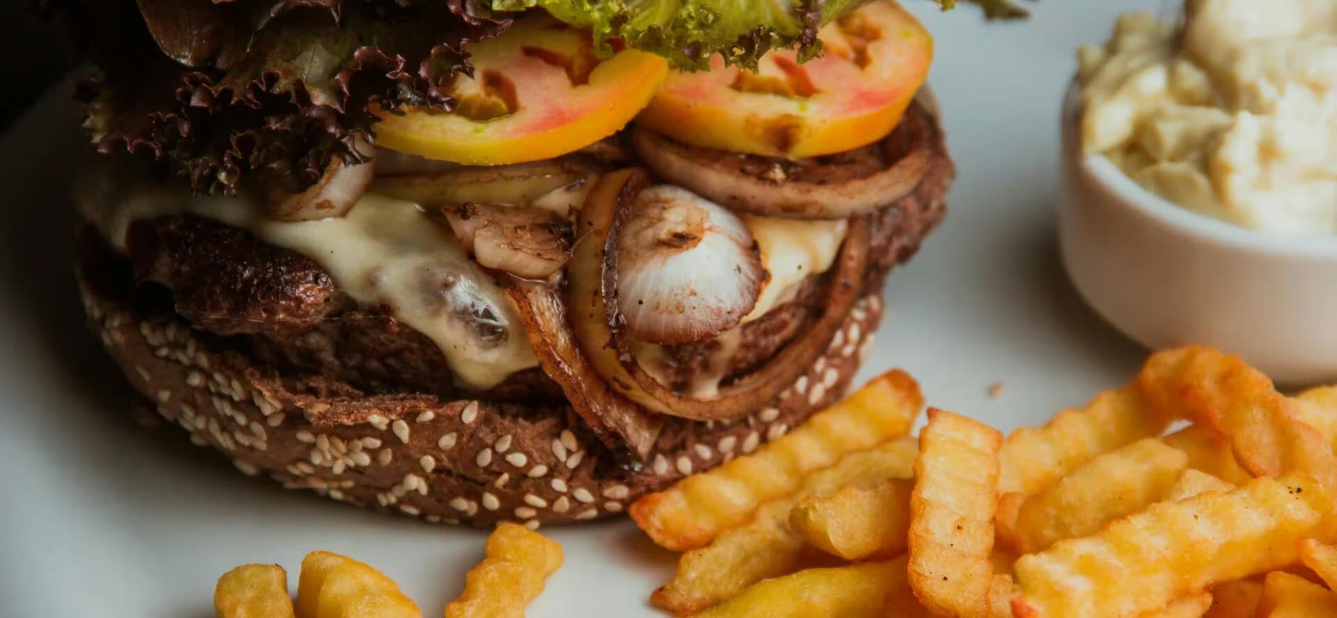 Een burger met rode sla, tomaat, gebakken ui en gesmolten kaas op een sesambroodje, met kroepoekfrites en een bakje saus ernaast.